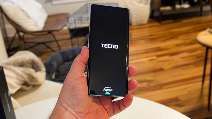 A person holding a TECNO smartphone displaying the startup screen with the TECNO logo, set in a cozy indoor environment with wooden flooring and soft furnishings in the background.