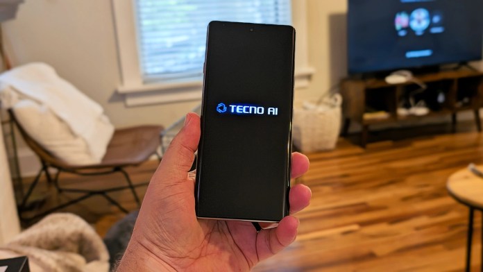A person holding the TECNO Camon 40 Pro 5G smartphone displaying the TECNO AI logo on the screen, in a cozy room setting with wooden flooring and a TV in the background.
