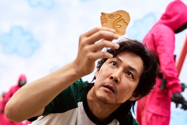 Close up of a man intensely examining a cookie while hooded guards in red walk around behind him; still from 