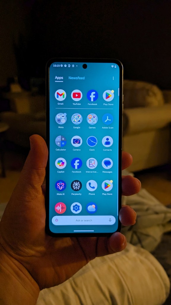 A person holding a smartphone displaying a user interface with various app icons and notifications, in a low-light room.