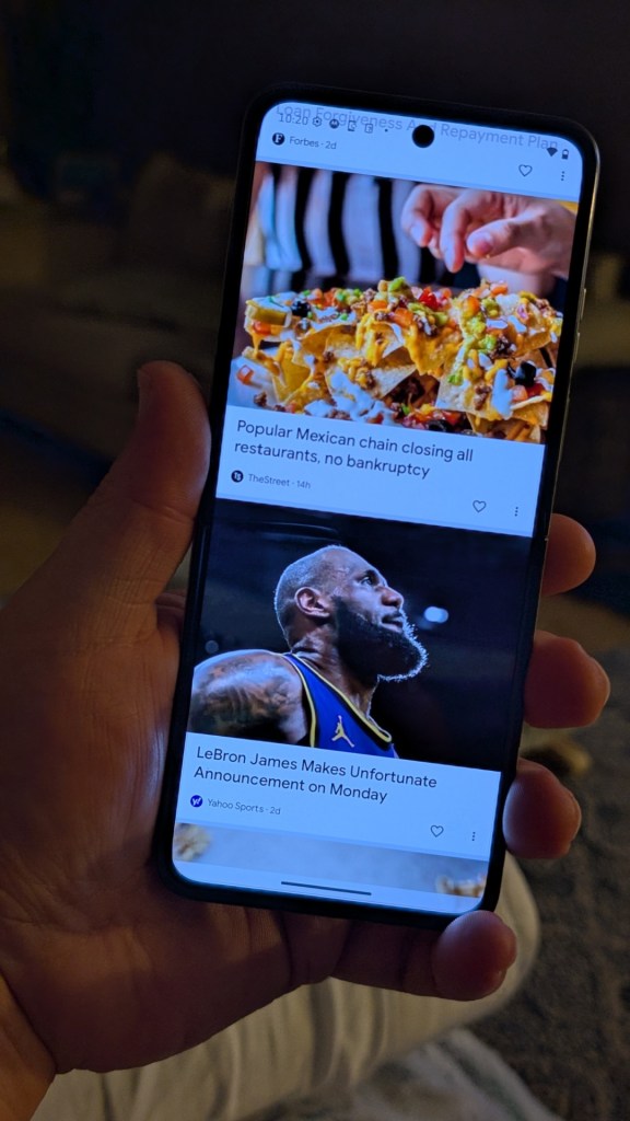A close-up of a hand holding a smartphone displaying a news feed, featuring various articles, including one about a popular Mexican chain closing restaurants and another about LeBron James.
