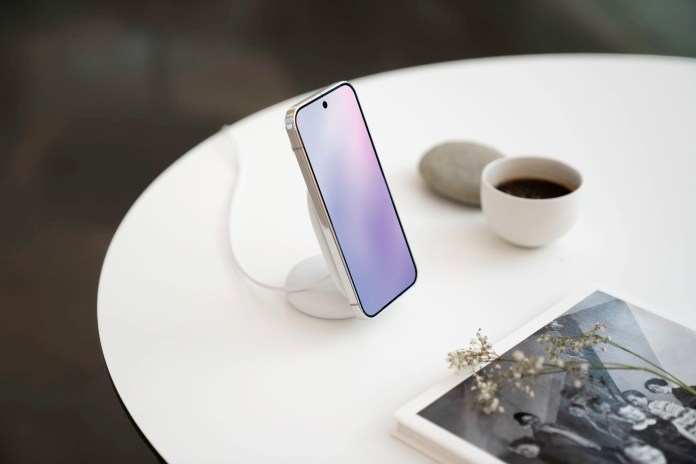 A smartphone placed on a wireless charging stand on a table, alongside a cup of coffee and a decorative stone.