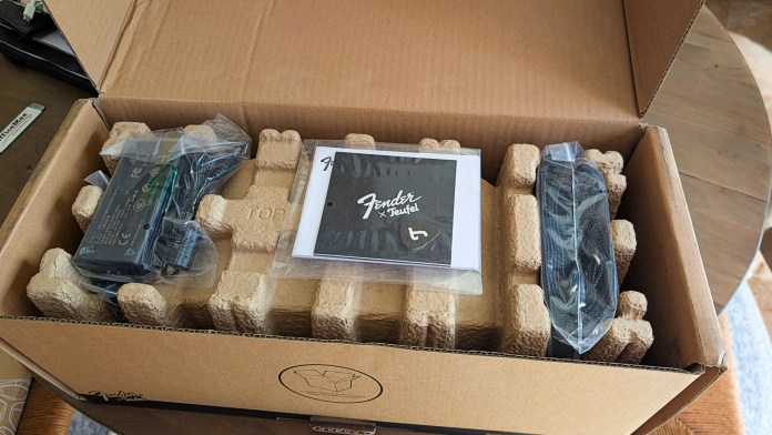 An open cardboard box containing the Fender x Teufel ROCKSTER CROSS speaker, power supply, and a booklet. The speaker is wrapped in plastic, alongside a Fender guitar pick.