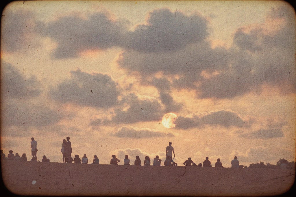 A line of people silhouetted along a ridge at sunset, standing and sitting beneath a clouded sky as the sun hangs low on the horizon.