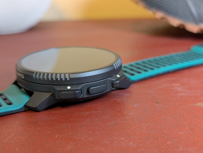 Close-up of a fitness smartwatch with a black face, side buttons, and a turquoise strap resting on a wooden surface.