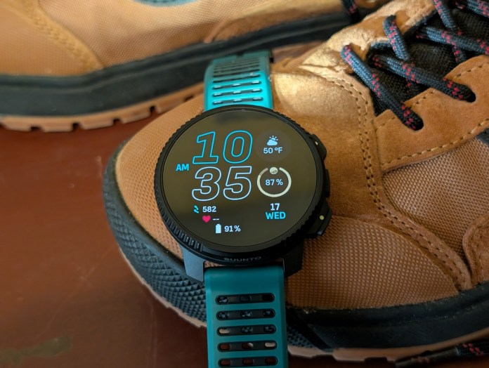 A Suunto smartwatch displaying the time, date, heart rate, and weather information, positioned next to a hiking shoe.